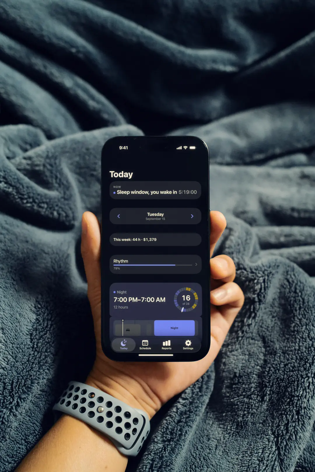 A hand holds an iPhone over a dark teal blanket in daylight. On the screen, TurnoRest’s Today at 9:41 on Tuesday, September 15: sleep window, you wake in 5:19:00; this week 44 h and $1,379; tonight a night shift, 7:00 PM to 7:00 AM.