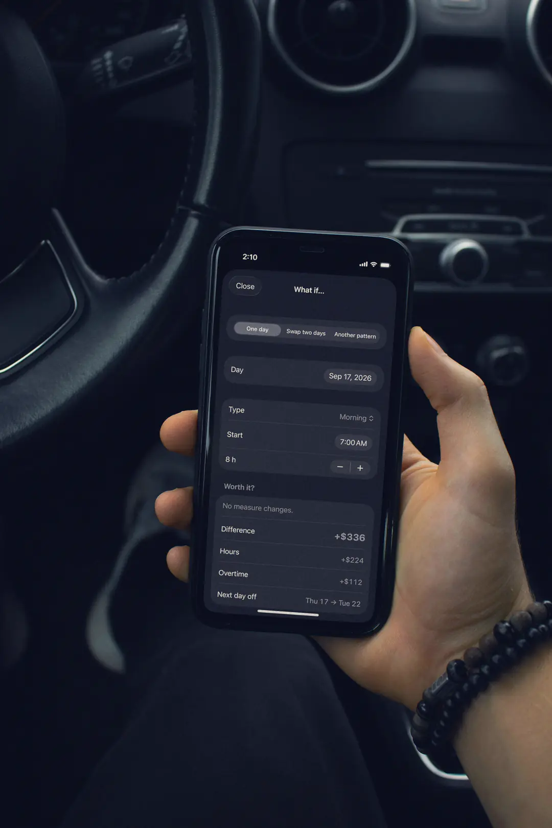 A hand holds an iPhone in a dark, parked car. On the screen, TurnoRest’s What if… sheet at 2:10: an 8-hour morning on Thursday, September 17, from 7:00 AM, worth +$336 (hours +$224, overtime +$112).