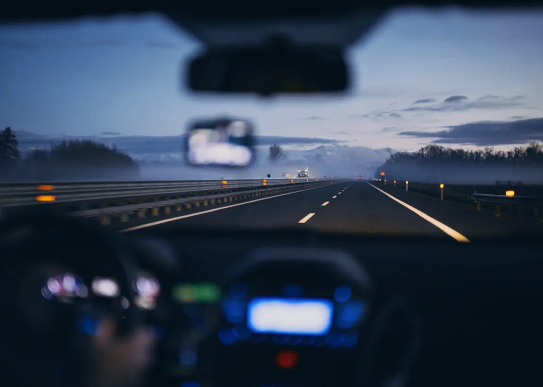 The view through a car windscreen onto a misty motorway at dawn, with the dashboard lights blurred in the foreground.