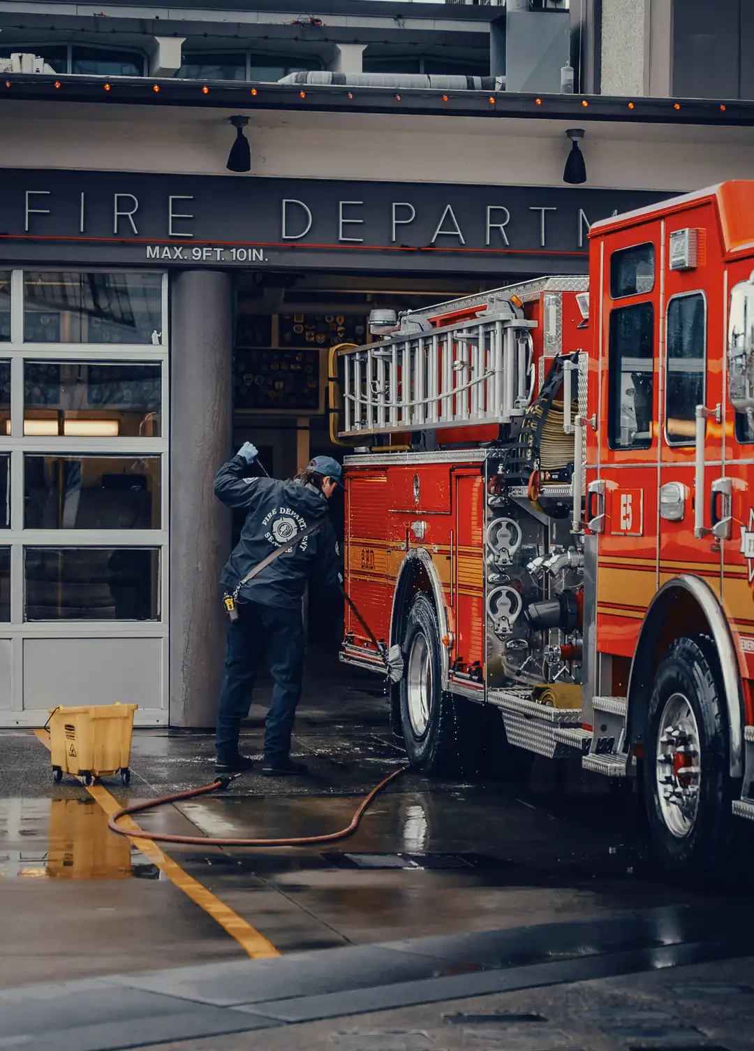 A firefighter in a navy jacket and cap scrubs a red fire engine with a long brush outside the open bay of a fire station, under the “FIRE DEPARTMENT” sign, with a hose and a yellow mop bucket on the wet apron.