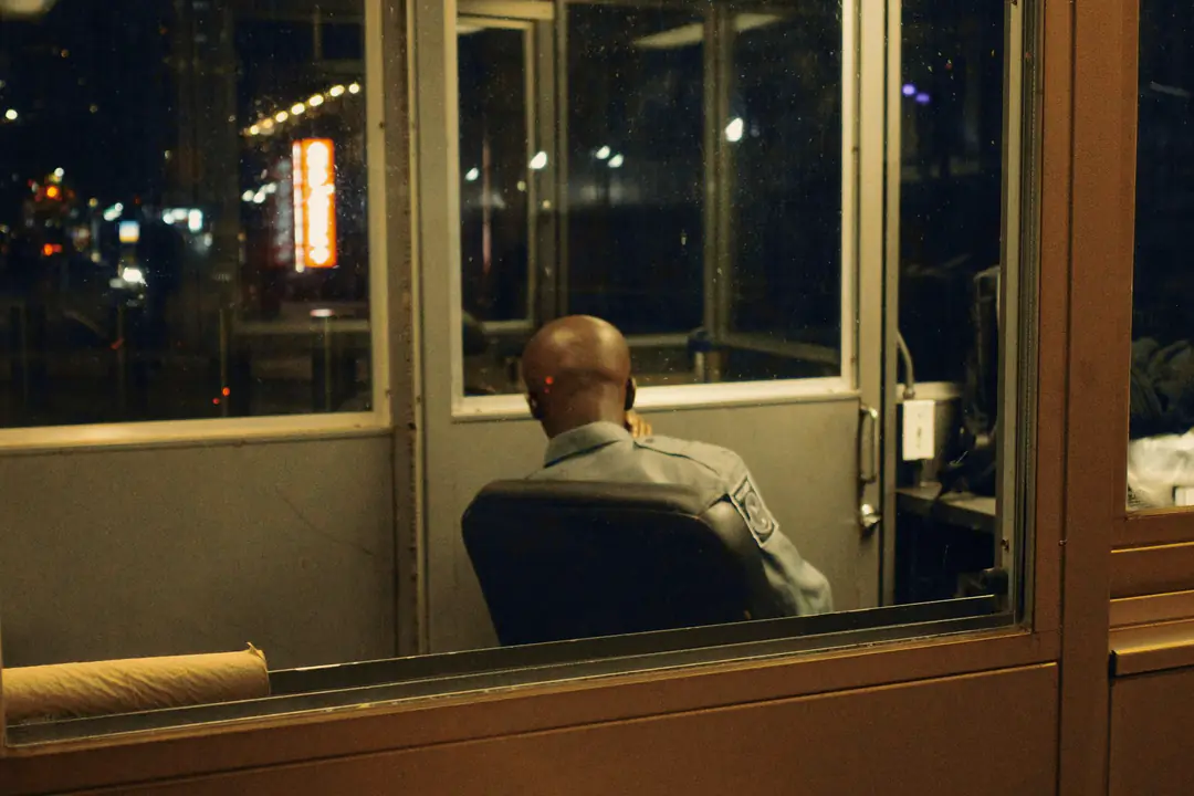 A security guard in a grey uniform sits alone in his lit booth at night, seen from behind through the glass, a hand at his ear, with the street lights blurred beyond the windows.