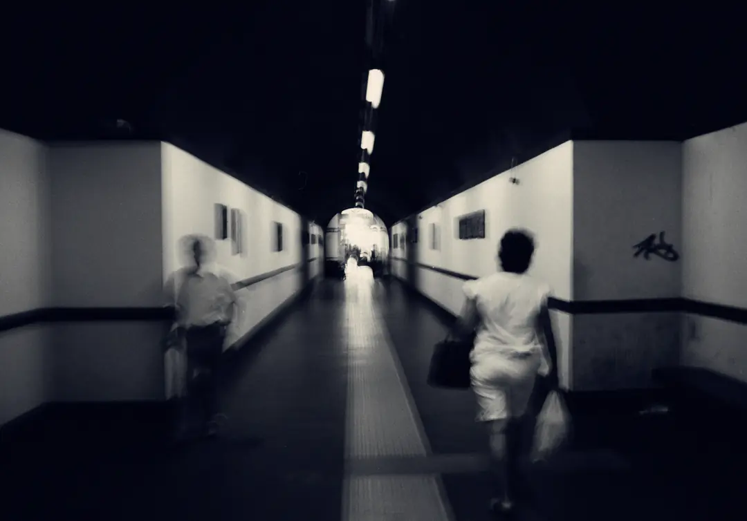 People walk through a long underground corridor under a row of ceiling lights towards the light at the far end, blurred by the long exposure. Black and white.