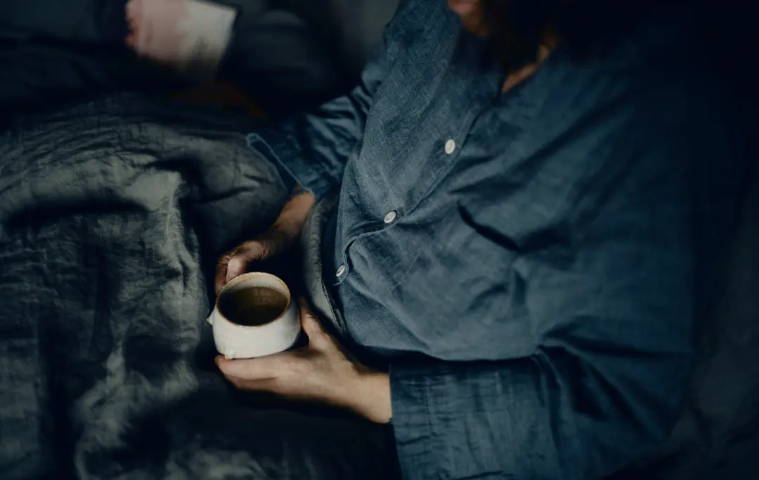 A person in a blue shirt sits in bed under a green duvet, holding a white mug of coffee in both hands. Their face is out of the frame.
