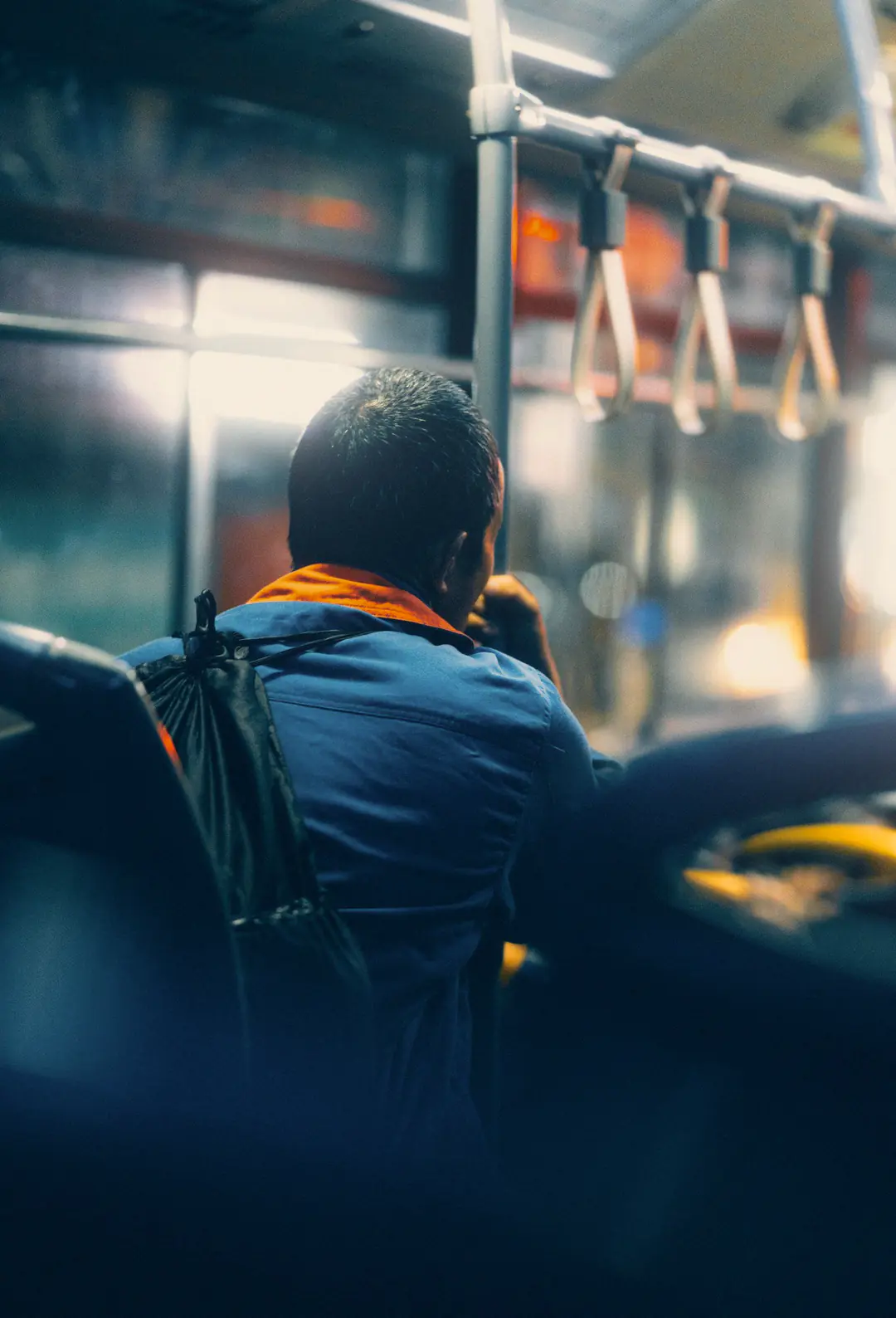 A man in a blue work jacket with a backpack, seen from behind, sits on a bus at night below the hand straps.