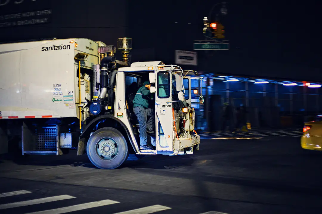 A sanitation worker in a green jacket climbs into the open cab of a white garbage truck as it turns a corner at night, with a yellow taxi and blue-lit scaffolding behind.