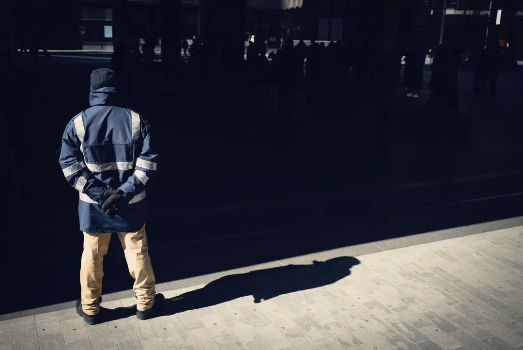 A guard in a navy jacket with reflective stripes stands with his hands behind his back on a sunlit pavement, seen from behind, his shadow long beside him.
