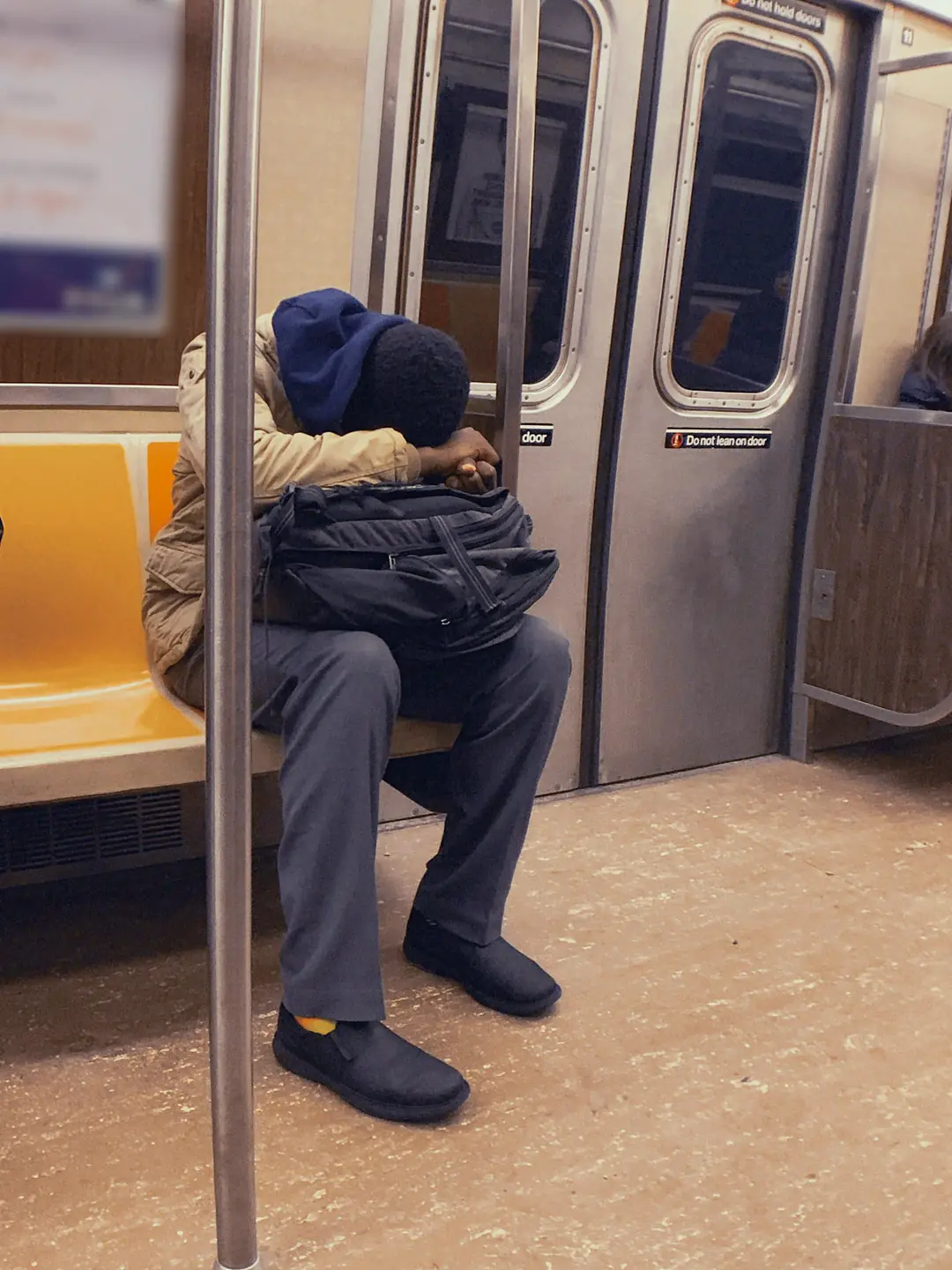 A man in a beige jacket, his hood up, sleeps folded over the backpack on his lap, on the orange seats of a subway car beside the doors.