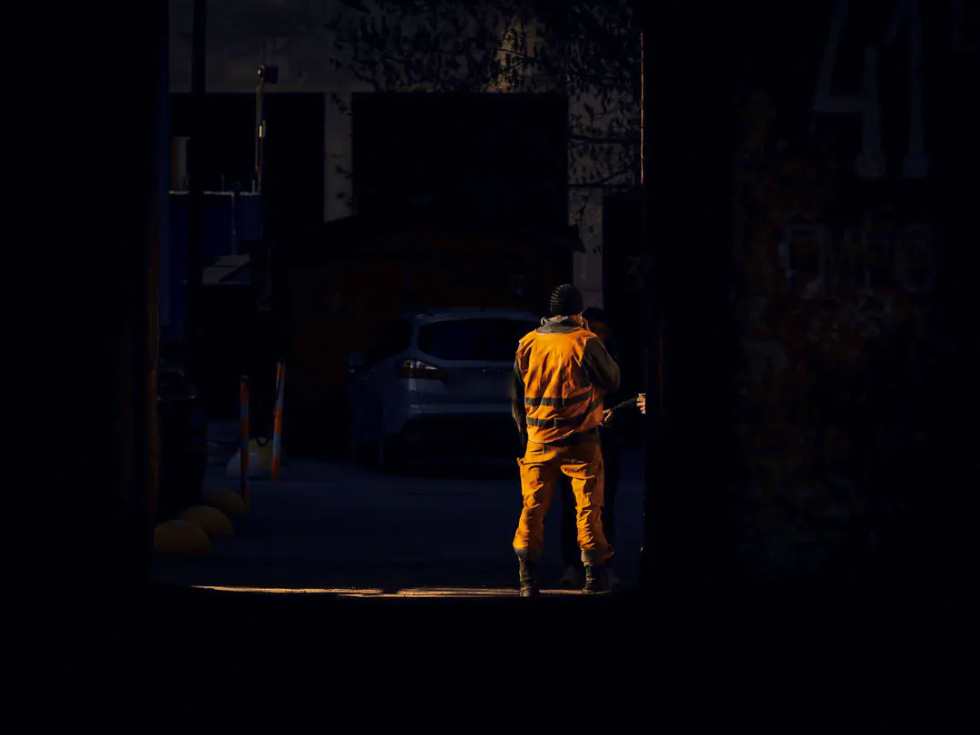 A worker in orange overalls and a reflective vest stands in a patch of light in a dark yard, with a parked car behind him.